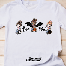Load image into Gallery viewer, The Boo-tiful, wheelchair Users shirt on a wooden table. A white t-shirt that says Boo-tiful! Three ladies loving the Halloween season. The first lady has dangling ghost earrings, and her wheelchair's wheels are black wheels with a ghost in the center. The lady in the middle is dressed as a bat, her bat wings are open, and her wheelchair's wheels have three bats shaping a circle and making a pumpkin in the middle. On her left, it says, "Boo," and on the right, it says, "tiful." The last lady has dangling sp
