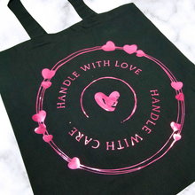 Load image into Gallery viewer, A Black tote bag with a circle rings frame with different hearts scattered throughout. Curved down, it says, &quot;Handle with Love.&quot; Curved up, it says, &quot;Handle with Care.&quot; In the middle is a damaged heart. 
