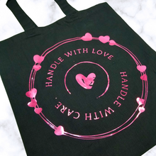 Load image into Gallery viewer, A Black tote bag with a circle rings frame with different hearts scattered throughout. Curved down, it says, "Handle with Love." Curved up, it says, "Handle with Care." In the middle is a damaged heart.