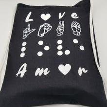 Load image into Gallery viewer, The black bag says "Love" in four languages. The first line is love in English. The letter "O" is replaced by a white heart. The second line has "love" in American Sign Language- fingerspelling. The third line has love in Braille. Finally, the four-line has "love" in Spanish - Amor. The letter "O" is replaced by a white heart. It has drawstring handles. The design uses raised vinyl except for "love" in American Sign Language- fingerspelling as the lines are too small to use raised vinyl.