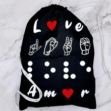 Load image into Gallery viewer, The backpack says "Love" in four different languages. The first line is love in English. The letter "O" is replaced by a red heart. The second line has love in American Sign Language- fingerspelling. The third line has love in Braille. Finally, the four-line has love in Spanish - Amor. The letter "O" is replaced by a red heart. It has drawstring handles.