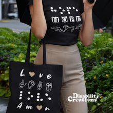 Load image into Gallery viewer, A woman is wearing a black shirt, the Hope in 3 languages tee with nude trousers holding an opened umbrella. She has the "Love in 4 Languages" with the nude hearts tote bag.
