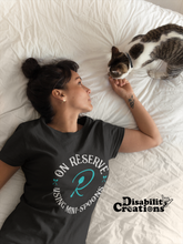 Load image into Gallery viewer, A woman with her hair up petting her cat in bed. She is wearing the On Reserve Using Mini-Spoons Shirt. The post says New Disability Shirt. July 28. www.DisabilityCreations.com
