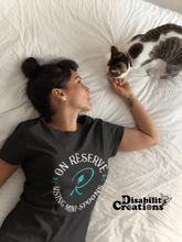 Load image into Gallery viewer, A woman with her hair up petting her cat in bed. She is wearing the On Reserve Using Mini-Spoons Shirt. The post says New Disability Shirt. July 28. www.DisabilityCreations.com