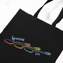 Load image into Gallery viewer, A Black tote bag with an outline of three engraved vintage spoons (pink, yellow, and turquoise) lined up side to side with the handles on the right. It says, &quot;Spoonie life.&quot; &quot;Spoonie&quot; is located above the first spoon. &quot;Life&quot; is below the third spoon&#39;s handle. 
