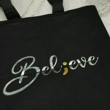 Load image into Gallery viewer, A tote bag that has "Believe" in silver handwringing with a semicolon replacing the "I."