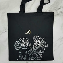 Load image into Gallery viewer, Tote bag that has a small gold butterfly is flying over silver line detail flowers. 

