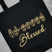 Load image into Gallery viewer, A Black tote bag says &quot;Blessed&quot; in two different languages in metallic gold vinyl. The first line is &quot;Blessed&quot; in American Sign Language-fingerspelling. The second line has &quot;Blessed in English. 
