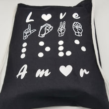 Load image into Gallery viewer, The black bag says &quot;Love&quot; in four languages. The first line is love in English. The letter &quot;O&quot; is replaced by a white heart. The second line has &quot;love&quot; in American Sign Language- fingerspelling. The third line has love in Braille. Finally, the four-line has &quot;love&quot; in Spanish - Amor. The letter &quot;O&quot; is replaced by a white heart. It has drawstring handles. The design uses raised vinyl except for &quot;love&quot; in American Sign Language- fingerspelling as the lines are too small to use raised vinyl. 

