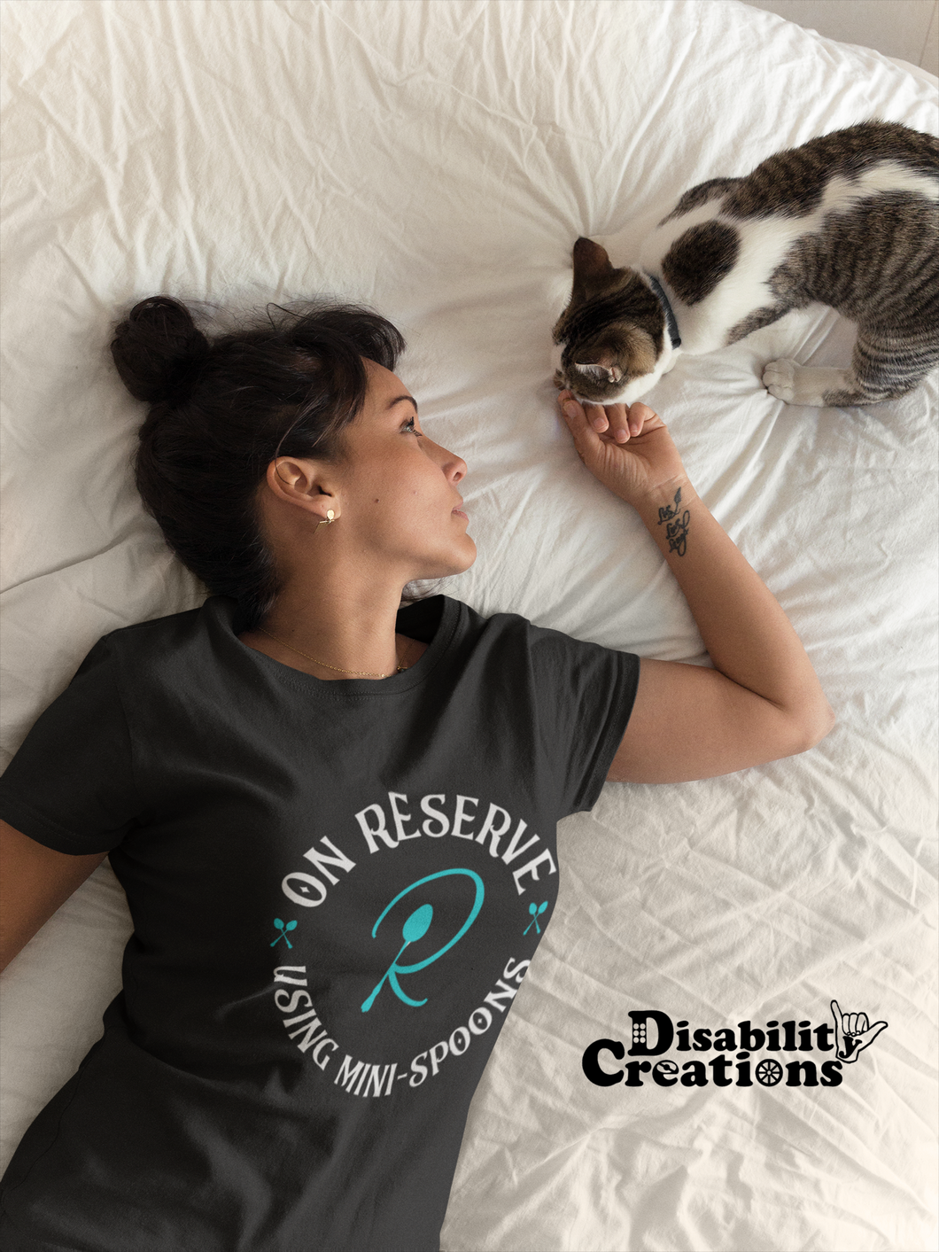 A woman with her hair up petting her cat in bed. She is wearing the On Reserve Using Mini-Spoons Shirt. The post says New Disability Shirt. July 28. www.DisabilityCreations.com