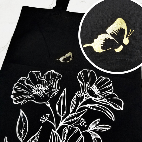 A Black double-sided tote bag. On one side, a small gold butterfly is flying over silver line detail flowers. The butterfly's body is a semicolon. On the other side, 