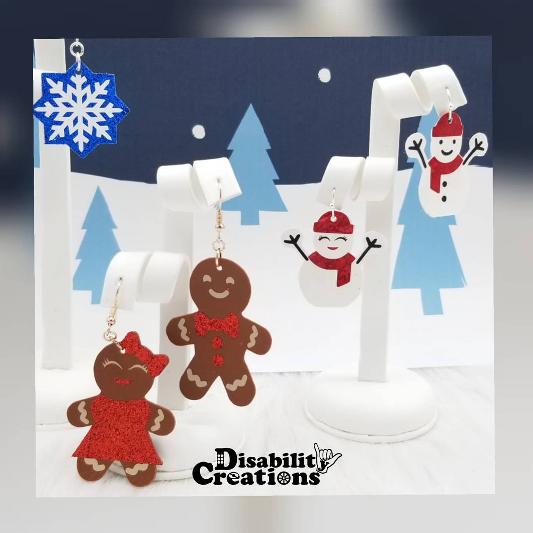 Mrs. &Mr. Snowie Earrings and the Gingerbread Couple Earrings hanging on earring stands with a Winter Wonderland background.