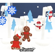 Load image into Gallery viewer, The Magical Snowflakes Earrings, Mrs. &amp;Mr. Snowie Earrings and the Gingerbread Couple Earrings hanging on earring stands with a Winter Wonderland background.
